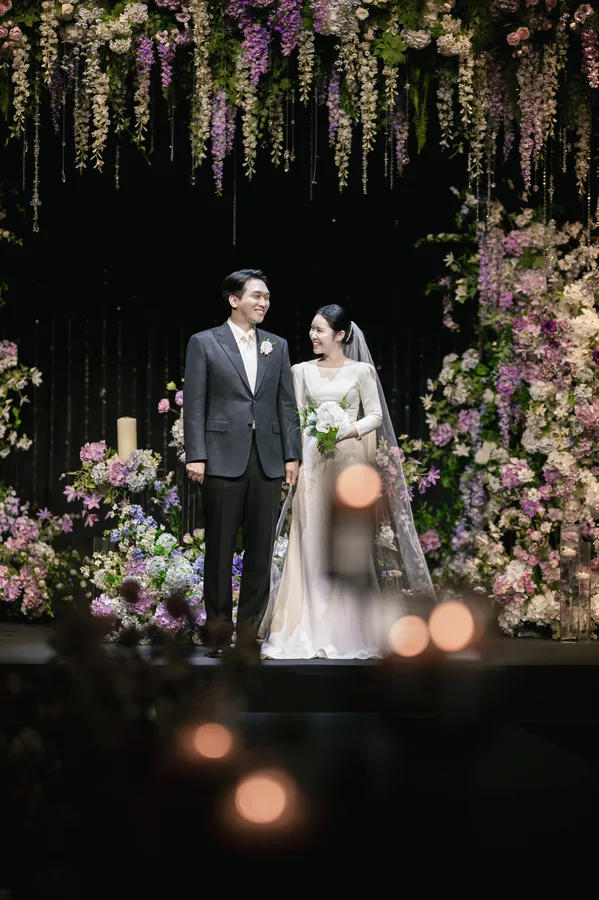 늘어진 등나무꽃 장식 아래 나란히 선 신랑신부와 흐린 촛불