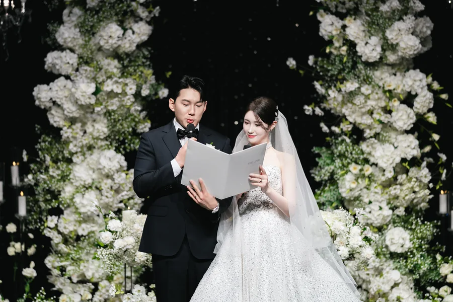 흰 꽃 아치 앞에서 성혼 서약서를 함께 읽는 신랑신부 클로즈업