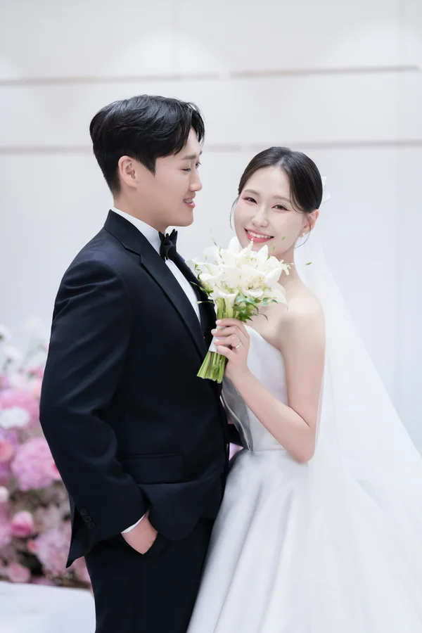 하얀 배경 앞에서 부케를 사이에 두고 마주보는 신랑신부의 다정한 옆모습