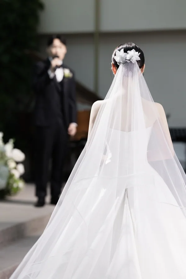 피아노 반주자를 배경으로 등을 보인 채 서 있는 신부 손에 흰 꽃 부케를 들었다