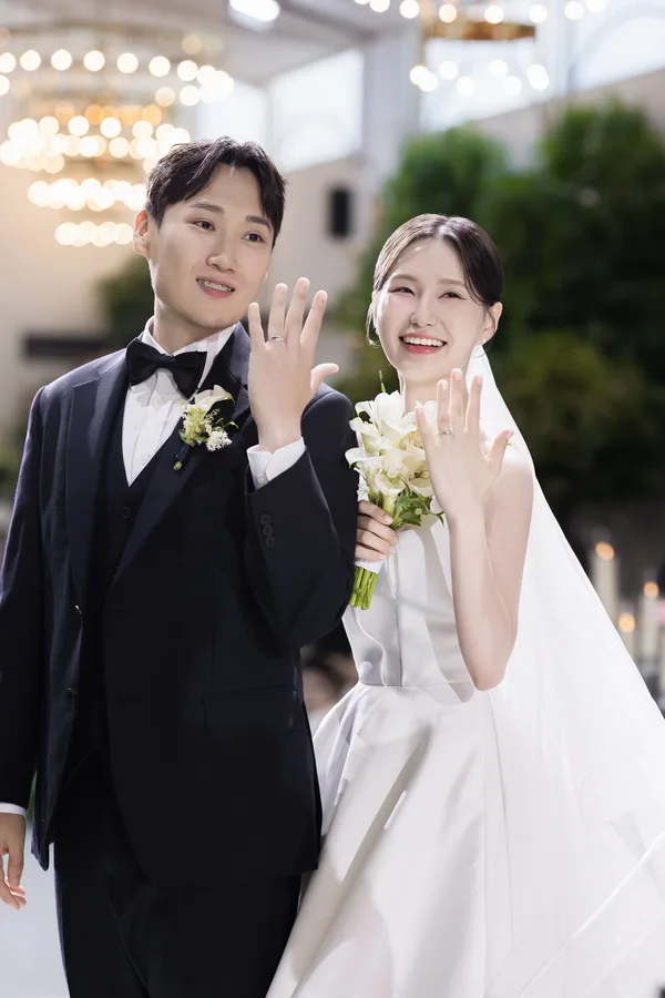 반지 낀 손을 나란히 들어 보이며 웃는 신랑신부 조명이 은은하게 배경을 비춘다