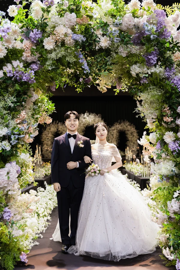 보랏빛 꽃 아치 입구에서 팔짱 끼고 선 신랑신부