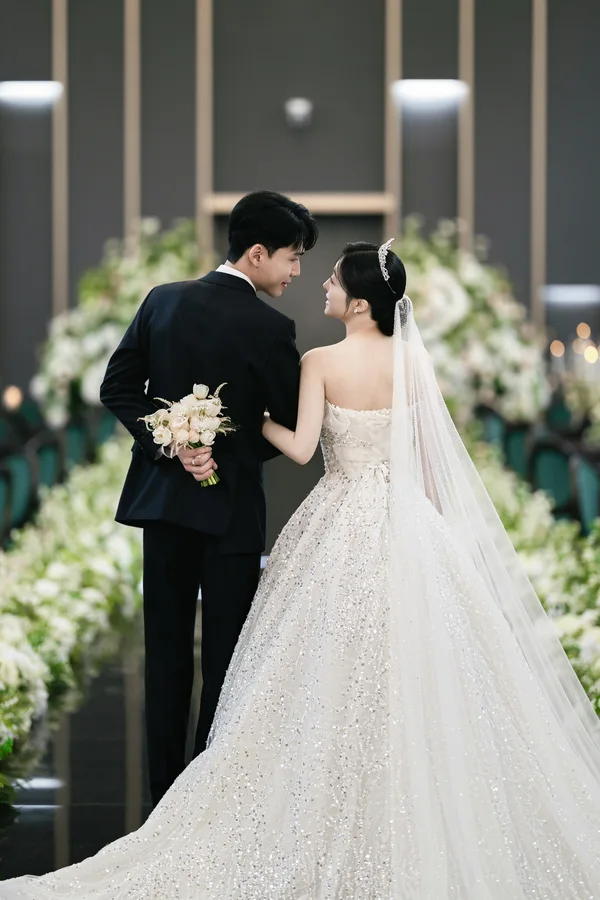 부케를 등 뒤로 감춘 신랑과 팔짱 낀 신부, 플라워로드를 걷는 뒷모습
