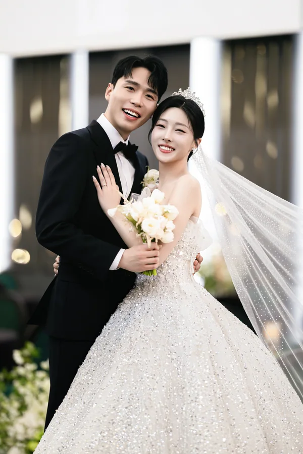밝은 기둥이 늘어선 배경 앞, 뺨을 맞대고 안은 신랑신부 클로즈업