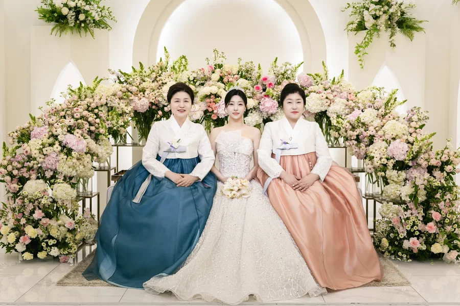 한복 입은 두 어머니와 함께 흰 아치 꽃장식 앞에 선 신부, 세 사람 기념사진